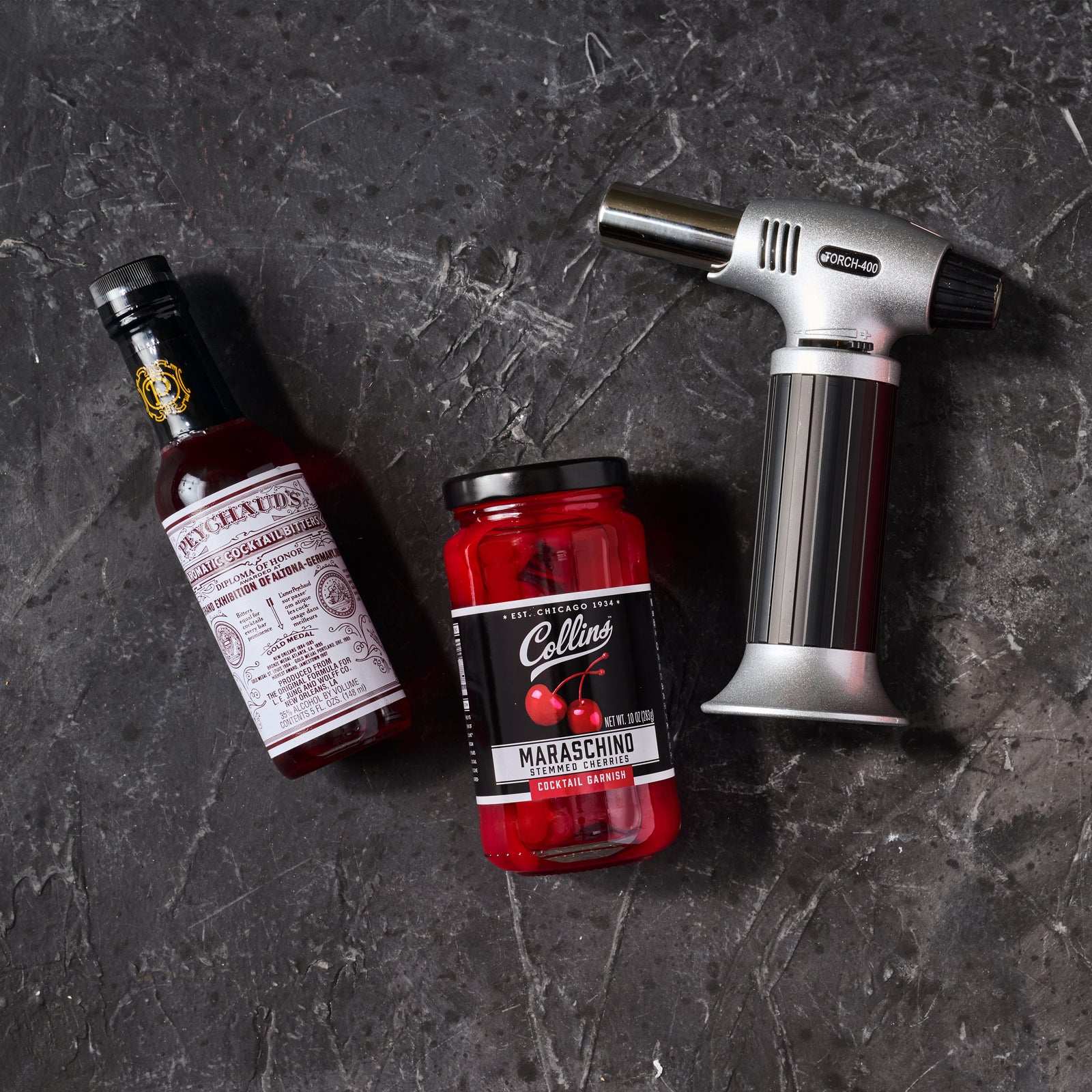 A butane torch, aromatic cocktail bitters, and a jar of Maraschino cherries one a slate background