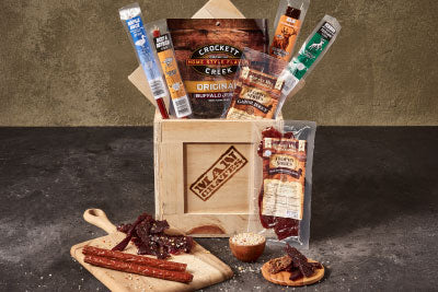 A Mancrates Mega Meat gift crate. There are many types of jerky emerging from a wooden crate, and more meats arranged on a wooden cutting board in front.