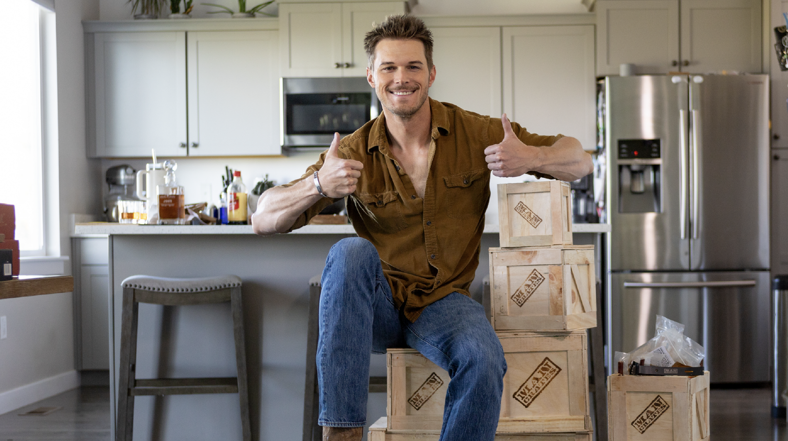  A rugged looking man sitting on a stack of wooden Mancrates gift crates giving two thumbs up in his kitchen. 