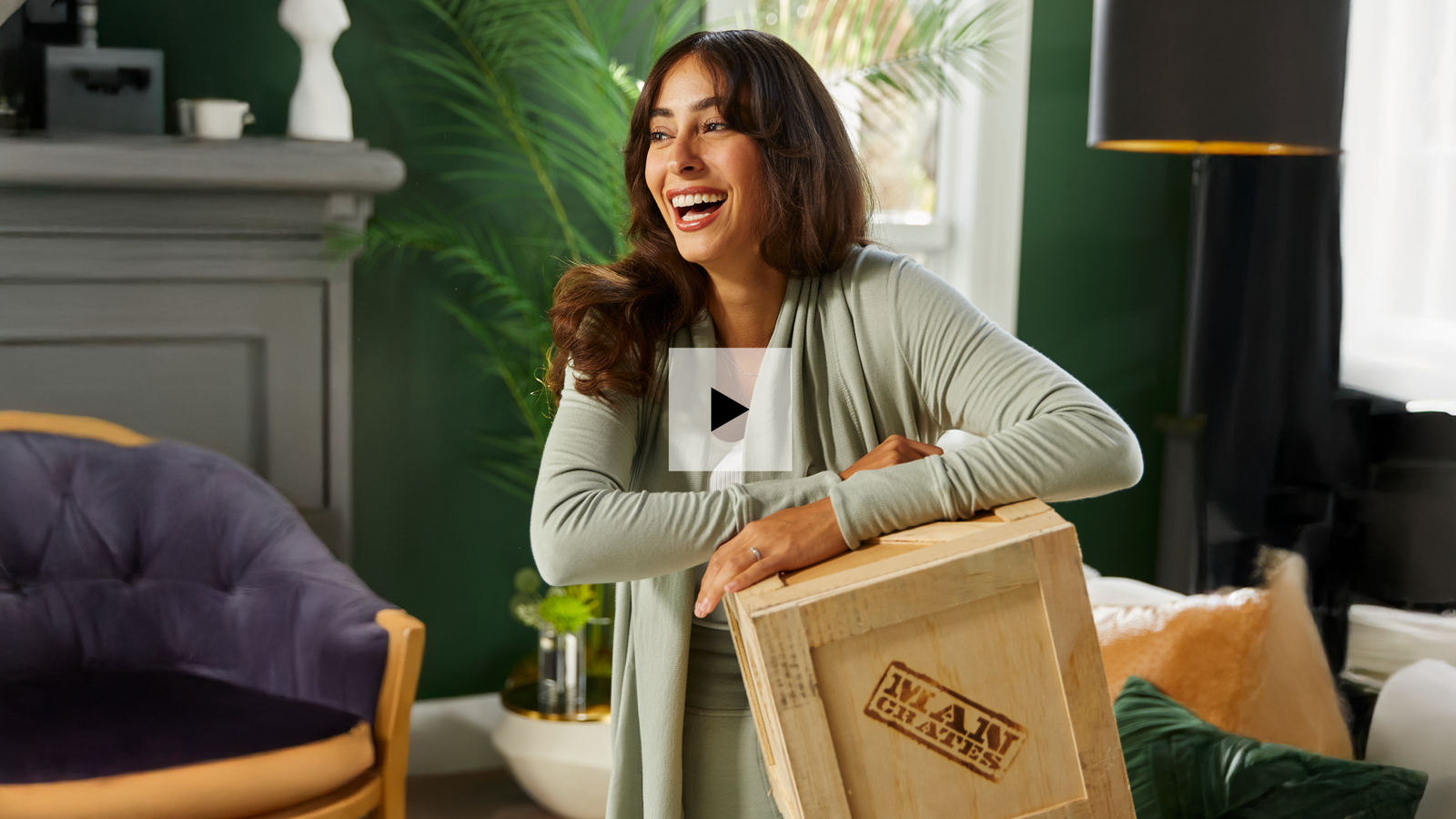 A beautiful woman smiling and holding a wooden Mancrates gift crate in a green room. There is a video play button superimposed on top of the image. 